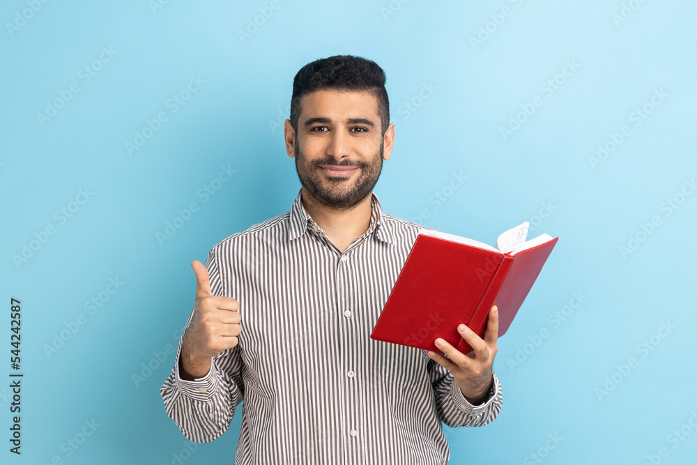 © khosrork - Portrait of smiling happy bearded businessman showing thumbs up gesture holding and reading book, likes genre and plot, wearing striped shirt. Indoor studio shot isolated on blue background.