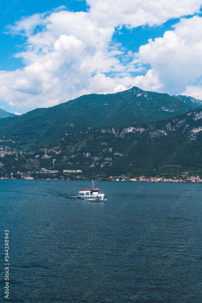 The boat ferry cruising along Lake Como near the shore of Varenna, a charming picturesque village in Lombardy, Italy, on a sunny summer day with the green Swiss mountains in the background. 