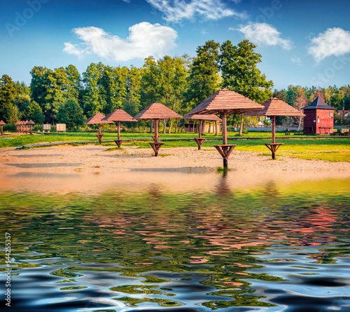 Wooden sun umbrellas on the empty beach reflected in the calm waters of Svityaz lake. Splendid morning scene on Shatsky National Park, Volyn region, Ukraine, Europe. Vacation concept background..