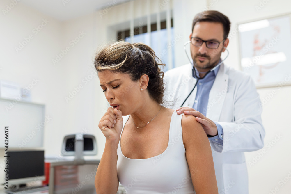 Obraz premium Young female patient in the clinic suffered from pneumonia, she is coughing the doctor listens to the wheezing in the lungs with a stethoscope.