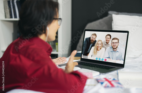 View from shoulder of mature woman in earphones using laptop to contact with group of people, video call with teammates from office, remote work concept