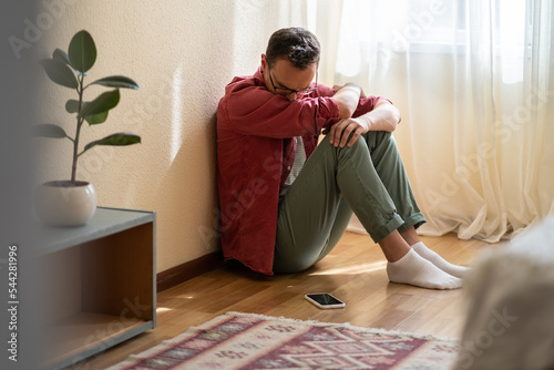 Depressed English man sits on floor of own apartment and sadly looks at mobile phone waiting for call. Disoriented casual guy stressed out because of being alone or breaking up with girlfriend