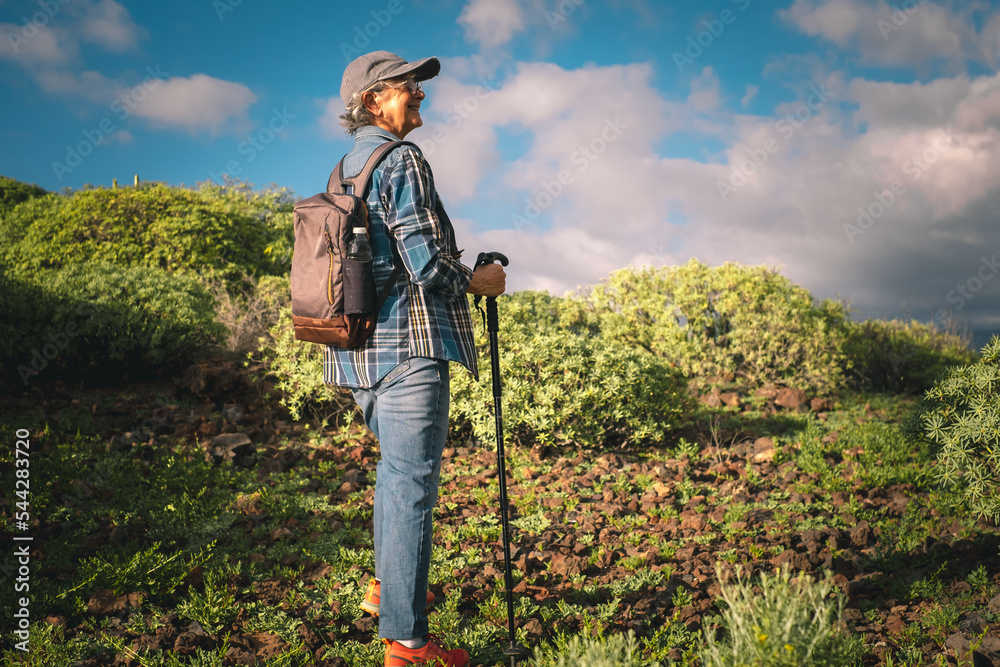 Portrait of active smiling senior woman enjoying trekking at sunset light. Elderly caucasian female with hat and backpack walking in mountain with a stick Portrait of active smiling senior woman enjoying trekking at sunset light. Elderly caucasian female with hat and backpack walking in mountain with a stick
