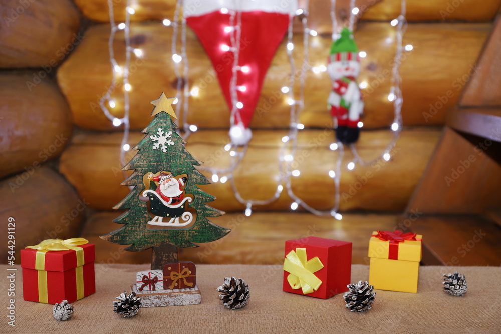 On a burlap-covered table are Christmas decorations and pine cones on the background of a garland and Santa hat hanging on a log wall.