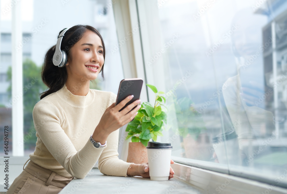 Gorgeous woman wearing headphone using mobile phone while standing near window in cozy room. Gorgeous woman wearing headphone using mobile phone while standing near window in cozy room.