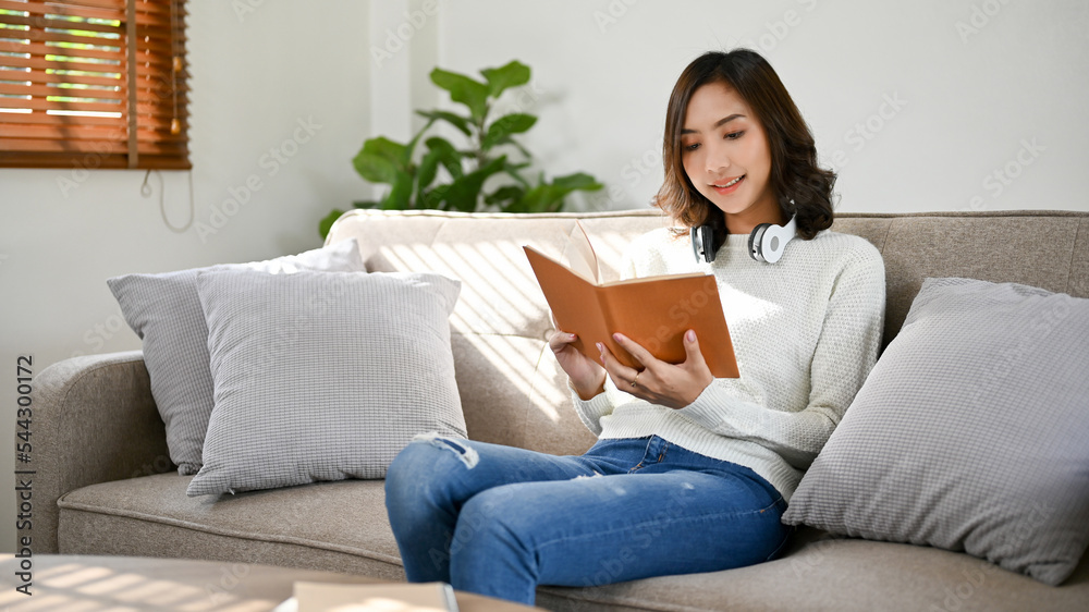 Gorgeous young Asian woman reading a book or novel while relaxing in her living room