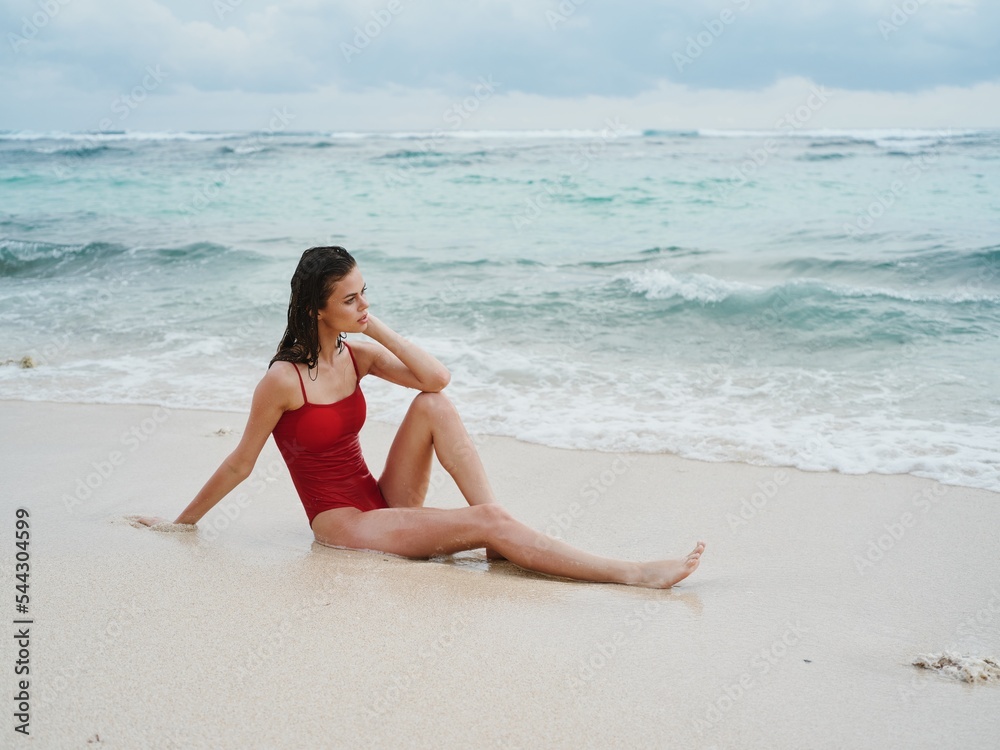 A woman in a red swimsuit sits on the sand by the ocean in the waves at Bali Island Beach and looks out at the beautiful view