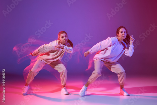 Portrait of two young girls dancing hip-hop isolated over gradient violet background in neon with mixed light