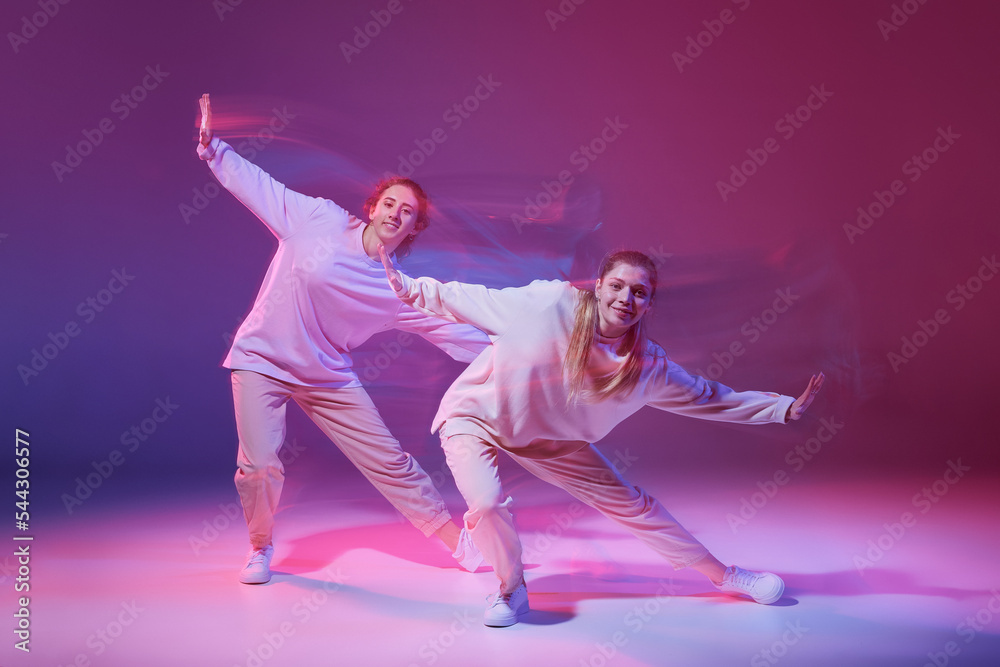 Portrait of young girls dancing hip-hop isolated over gradient purple background in neon with mixed light. Coordination of movements