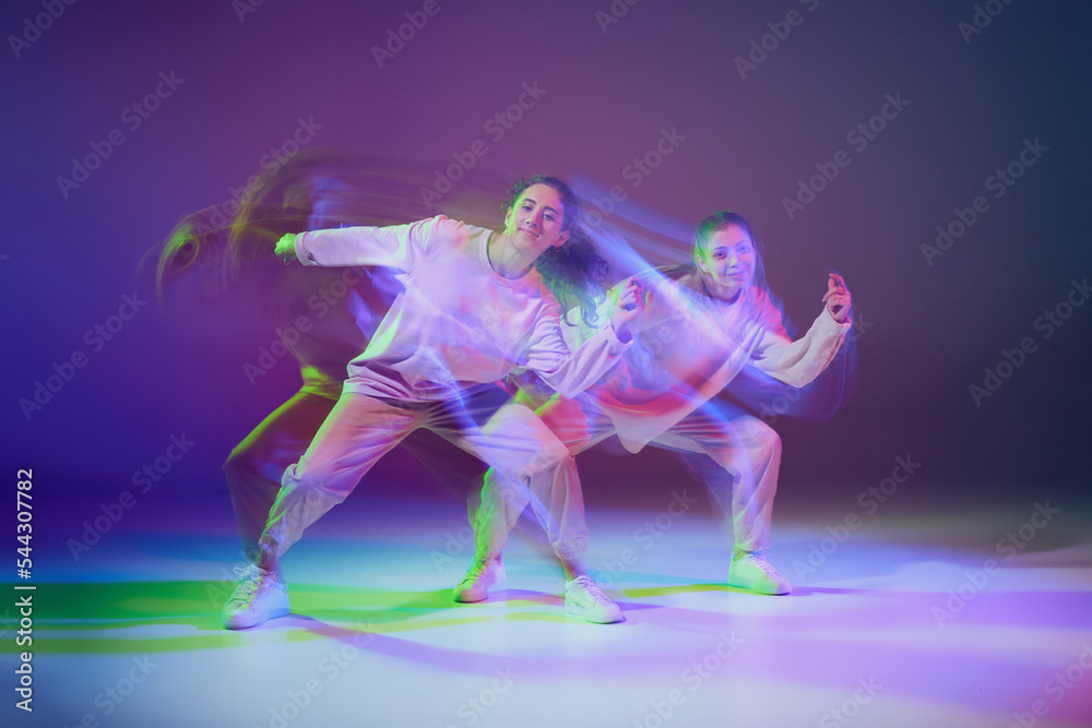Obraz premium Portrait of young girls dancing hip-hop isolated over gradient blue purple background in neon with mixed light. Energy burst