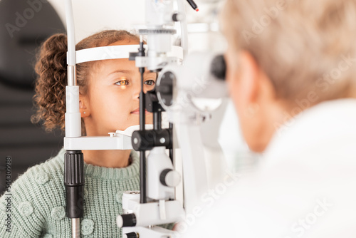 Child, kid or girl in optometry eye exam with optometrist, ophthalmologist or consulting medical profession and clinic slit lamp. Eye test, children eye care or vision check for healthcare insurance