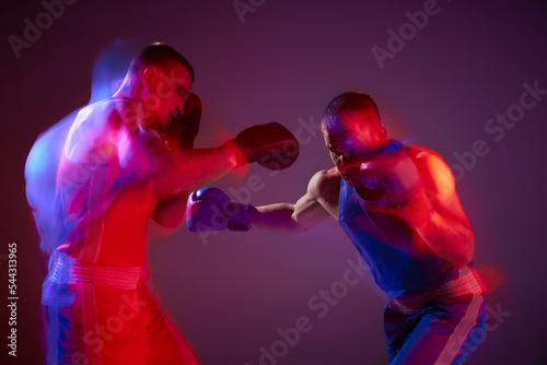 Unfocused effect portrait of two professional boxers boxing isolated on purple background in neon light. Concept of sport, competition, training, energy. © master1305