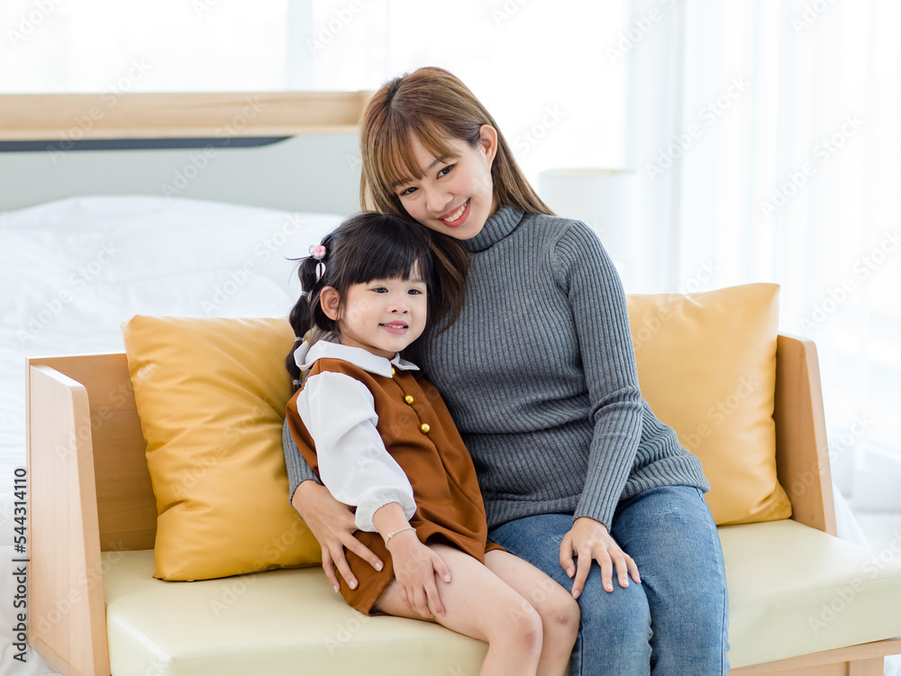 Millennial Asian cheerful happy young pretty female teenager mother nanny babysitter in casual outfit sitting on cozy sofa smiling hugging showing love with little cute preschooler daughter girl