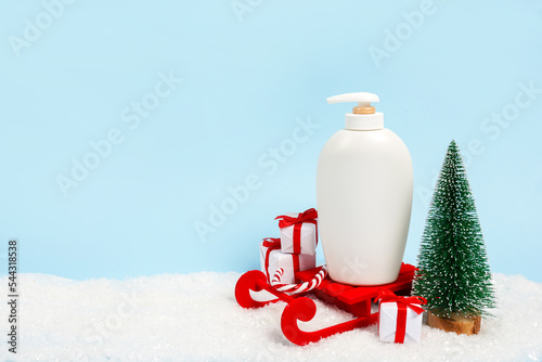 Cosmetic white bottle staying on red sled near christmas tree on blue background. Christmas and New Year sale, gifts, beauty and skin care concept