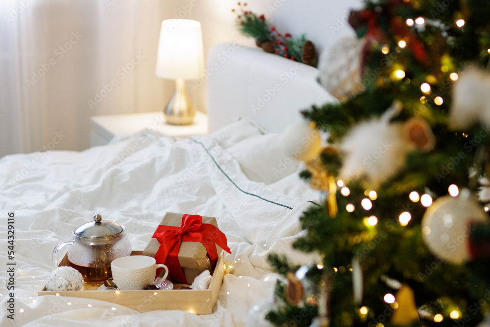Christmas concept - wooden tray with coffee or tea mug and gift box in decorated bedroom with Christmas tree Christmas concept - wooden tray with coffee or tea mug and gift box in decorated bedroom with Christmas tree