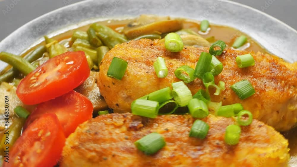Chicken cutlets with green beans and champignons in tomato sauce garnished with tomatoes and green onions in a plate rotates in a circle. Close-up Chicken cutlets with green beans and champignons in tomato sauce garnished with tomatoes and green onions in a plate rotates in a circle. Close-up