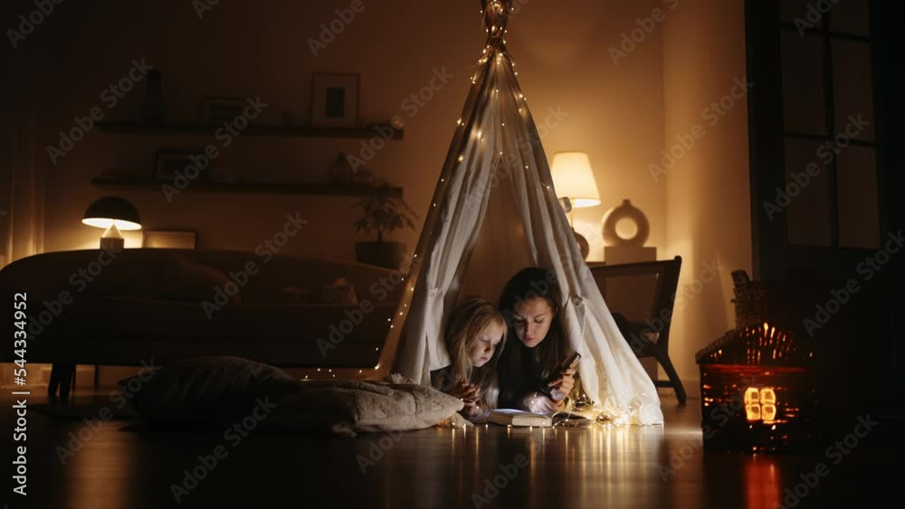 happy little girl and her mom are playing in teepee tent in room in evening, reading book