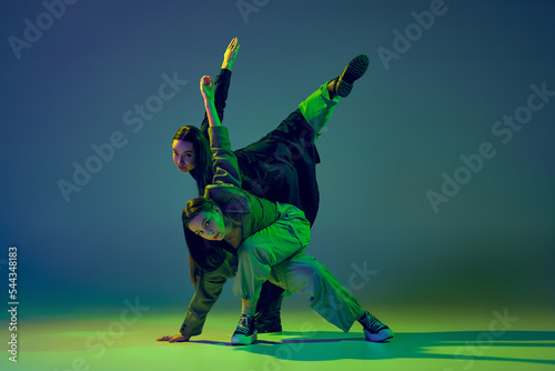 Energy. Stylish young girls dancing casual clothes dancing experimental style dance over gradient blue-green background at dance hall in neon light. Youth culture photo