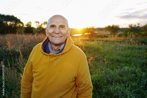 Elder man wearing yellow hoodie posing to camera outdoor. Portrait of mature happy farmer smiling with teeth.