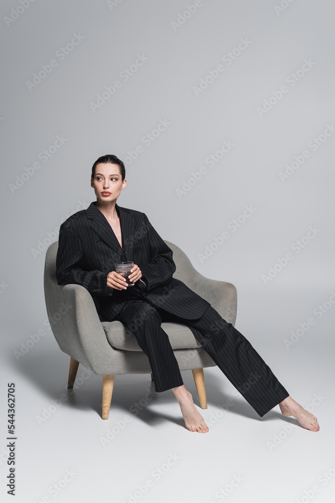 Barefoot woman in black suit holding whiskey glass on armchair on grey background. Barefoot woman in black suit holding whiskey glass on armchair on grey background.