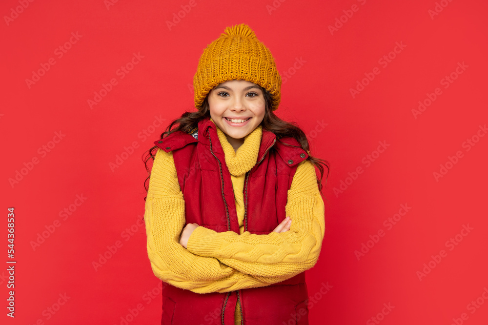 smiling child in knitted winter hat and down vest on red background, cold season