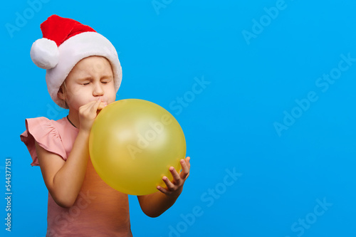 Happy new year concept. Caucasian preschool girl wearing santa claus red hat and glasses inflate yellow balloon on blue isolated background