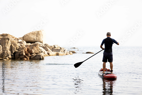 Senior man paddle surf on the beach. Male over 50 aging youthfully, paddling and doing exercise at early morning. Older adult adventurer having fun and enjoying. Peaceful and relax. Copy space. photo
