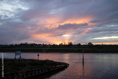 Dramatic sunset sun rays over river IJssel with silhouetted trees of vibrant intense color sunset sky with texture and detail of cloud cover lighting up orange, red and magenta tones. © Maarten Zeehandelaar