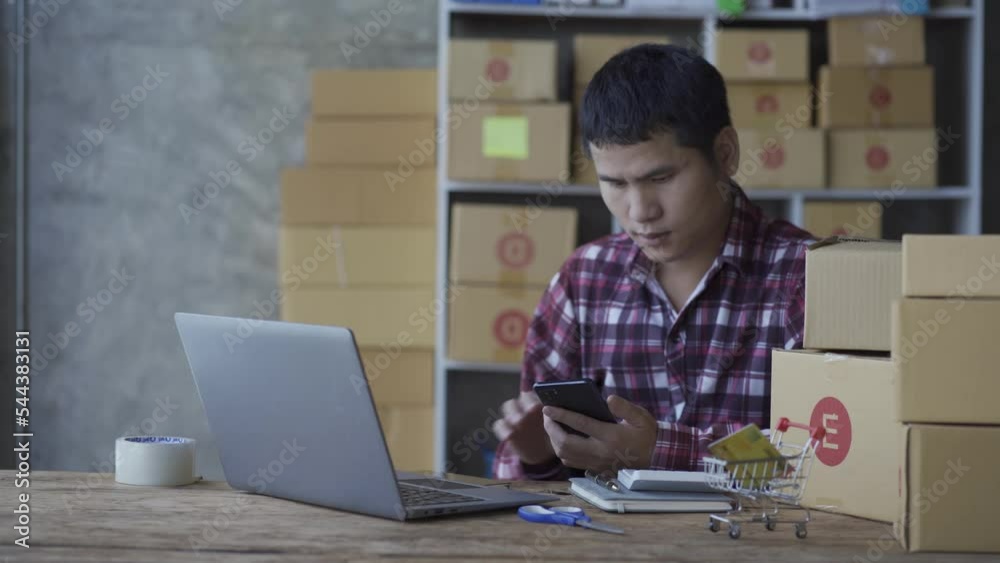 Young man talking on smartphone and using laptop computer to sell products online at home. Sales person packing products into boxes to deliver to customers who order on online store sites. Seller, sho Young man talking on smartphone and using laptop computer to sell products online at home. Sales person packing products into boxes to deliver to customers who order on online store sites. Seller, sho
