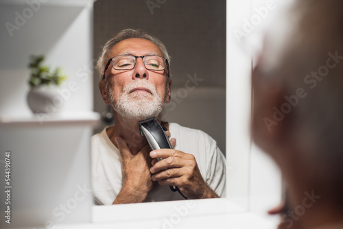 Senior man is trimming his beard with electric trimmer in bathroom.
