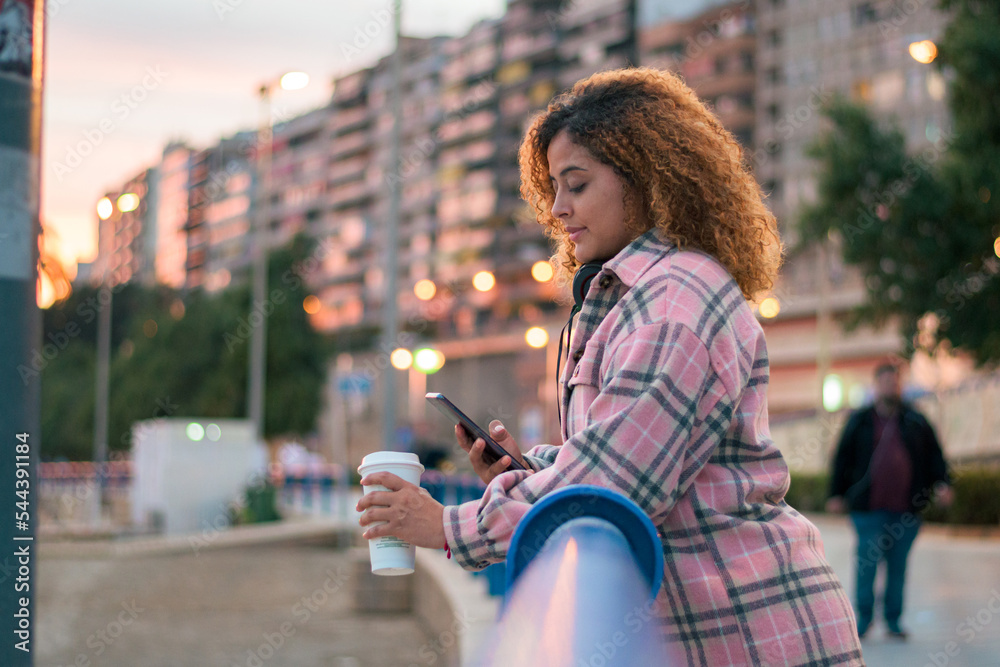 Fototapeta premium Curvy, curly-haired woman looks at her phone while drinking coffee on the street
