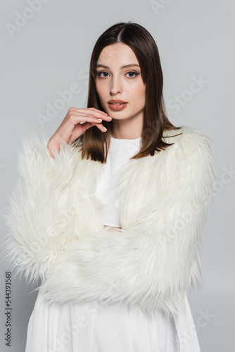 portrait of pretty young woman in stylish white faux fur jacket looking at camera isolated on grey.
