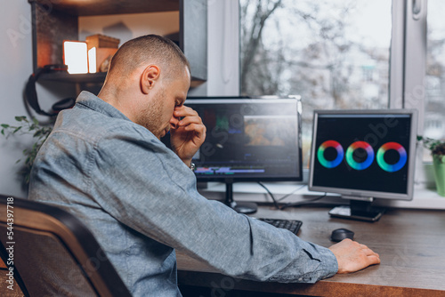 Side view of exhausted male freelancer rubbing eyes and  having headache during work at table with computer screen in home studio 