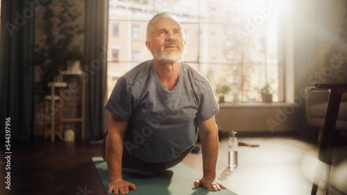 Portrait of a Strong Fit Senior Man Training on a Yoga Mat, Doing Back Stretching and Core Strengthening Exercises During Morning Workout at Home in Sunny Apartment. Concept of Health and Fitness.