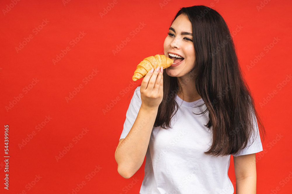 Portrait of young beautiful hungry woman eating croissant. Isolated portrait of woman with fast food over red background. Diet breakfast concept. Portrait of young beautiful hungry woman eating croissant. Isolated portrait of woman with fast food over red background. Diet breakfast concept.