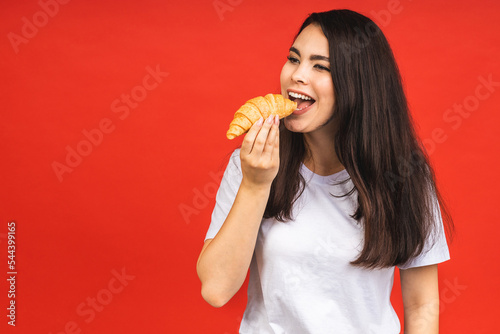 Portrait of young beautiful hungry woman eating croissant. Isolated portrait of woman with fast food over red background. Diet breakfast concept.