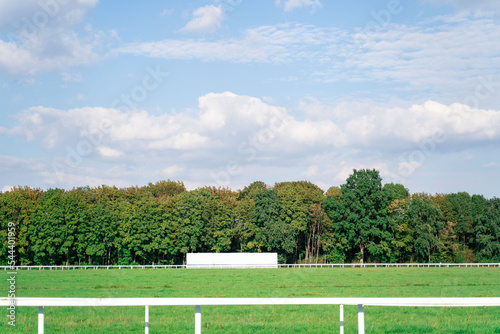 Empty high-speed track with partitions at hippodrome for horse racing surrounded by dense green forest with trees and cloudy blue sky on background. Gambling, betting, weekend activity, holiday events