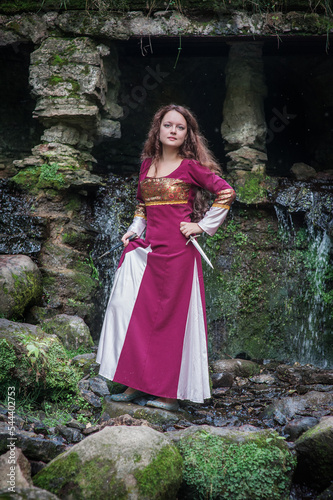 Young beautiful woman wearing medieval style dress with dagger near waterfall