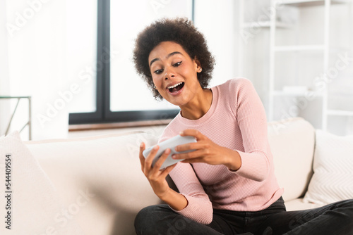 Happy millennial african american female with open mouth playing online game on phone, sits on sofa