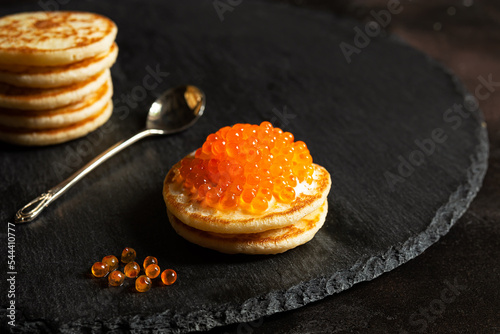 Red caviar on small pancakes blinis with sour cream and a spoon on a slate