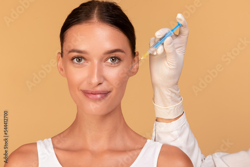 Happy young beautiful woman getting facial injection in cheekbones zone, standing over beige studio background photo