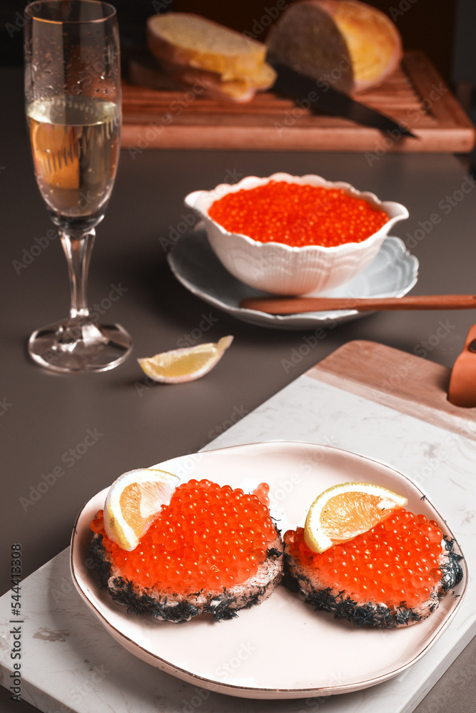 two sandwiches with red fish caviar served with a glass of champagne and a slice of lemon, in the background a porcelain white bowl filled with caviar with a teaspoon and sliced wheat bread
