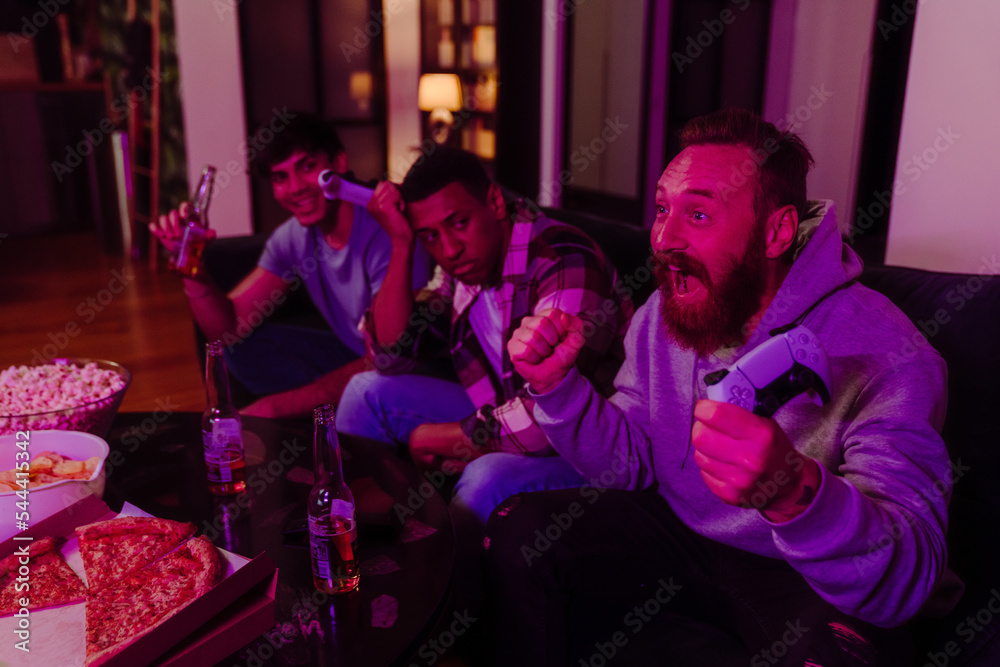 Three male friends playing video games and drinking beer in dark living room