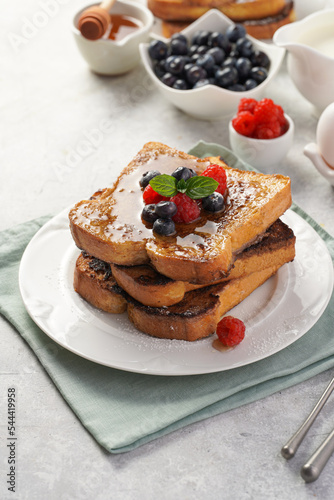 Several pieces of french toast - white wheat bread soaked in egg  milk and sugar  fried on a pan - stacked on white plate with fresh raspberries  blueberries  honey  mint on light grey background