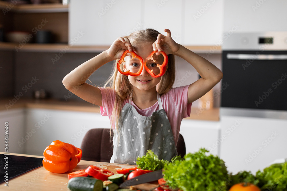 Cheerful european little kid puts pieces of pepper to her eyes, has fun in modern kitchen interior Cheerful european little kid puts pieces of pepper to her eyes, has fun in modern kitchen interior