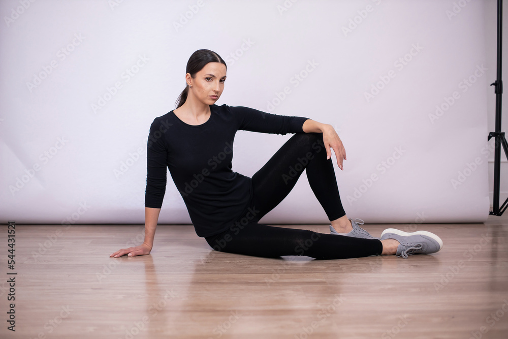 full length woman sit and show poses on the floor in black tight fitting clothes and sneakers in the studio on a white background.