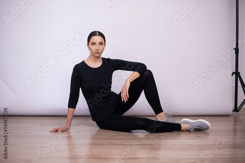 full length woman sit and show poses on the floor in black tight fitting clothes and sneakers in the studio on a white background.