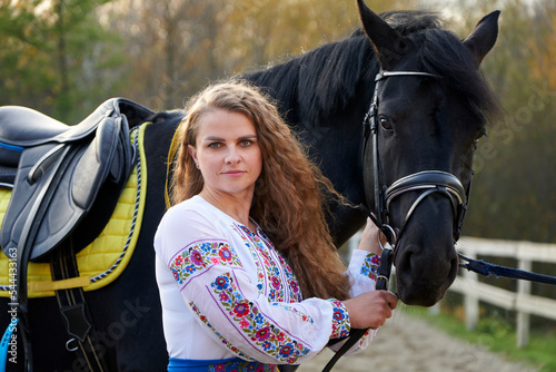 Young sexy woman standing near a horse in authentic suit. Beautiful professional female jockey standing near horse. A cute female caresses her favorite horse. Golden hour.