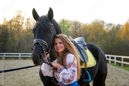 Young sexy woman standing near a horse in authentic suit. Beautiful professional female jockey standing near horse. A cute female caresses her favorite horse. Golden hour.