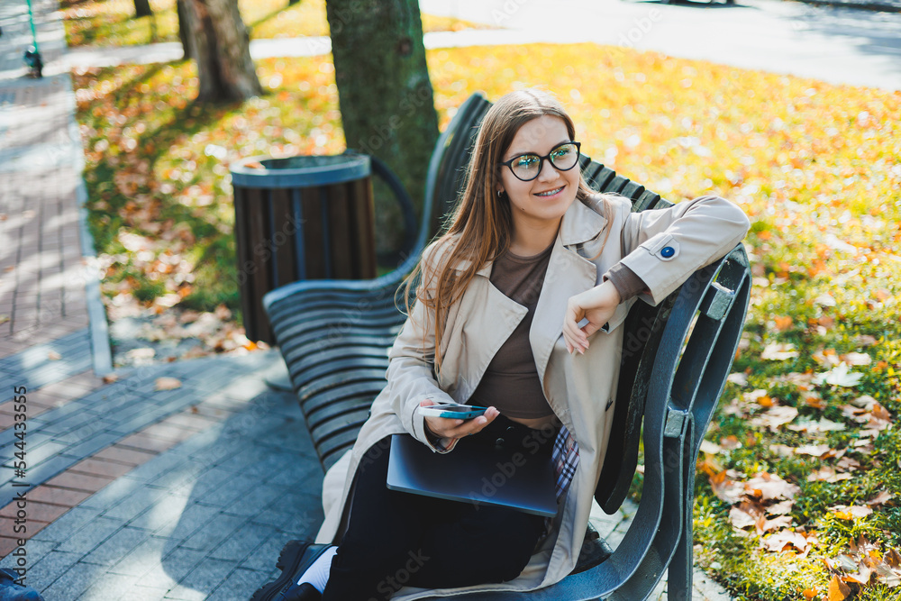 A smiling woman works with a laptop on a bench in the Autumn Park. She has a great smile, long hair and big blue eyes. Portrait of a modern working woman. Yellow park background A smiling woman works with a laptop on a bench in the Autumn Park. She has a great smile, long hair and big blue eyes. Portrait of a modern working woman. Yellow park background